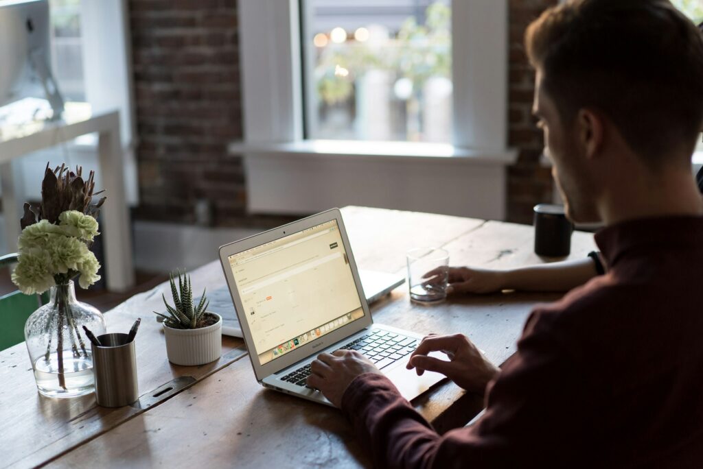 Over-the-shoulder view of a man sitting at a desk in an office, who appears to be working on his laptop. On the desk is a potted plant, a vase of flowers, and a few cups. There is a window in the background that lets in the light. There appears to be someone next to the man.